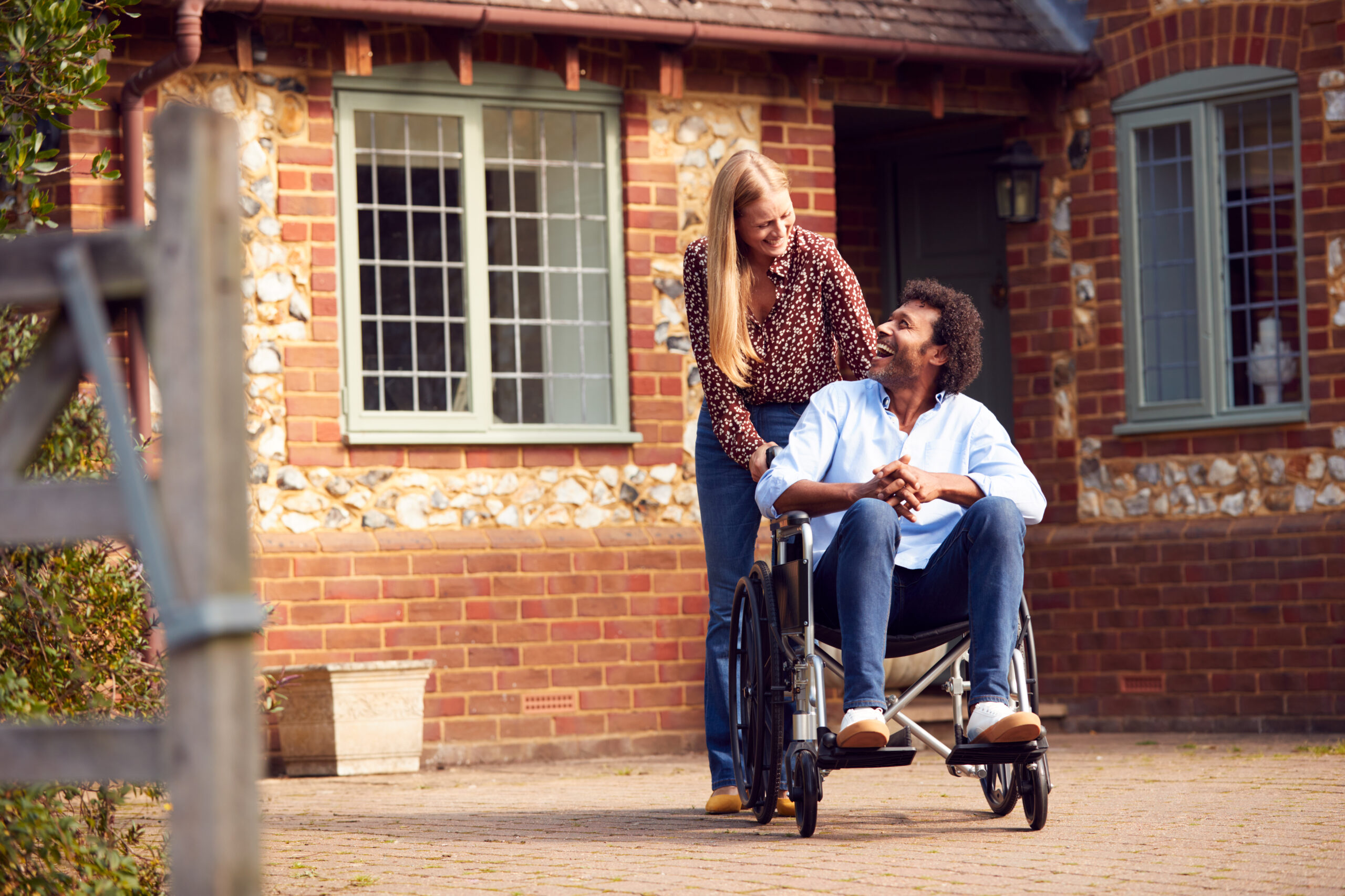 Mature Couple With Man Sitting In Wheelchair Being Pushed By Woman Outside Home